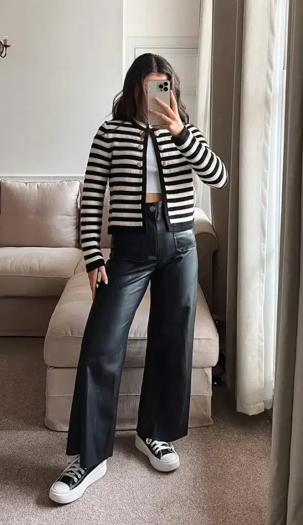 A woman taking a mirror selfie in a neutral living room, wearing a black and white horizontal-stripe cropped knit jacket with gold button closure over a white fitted crop top, paired with high-waisted wide-leg faux leather black trousers, and black and white platform canvas trainers, with a cream sofa and armchair visible in the background.