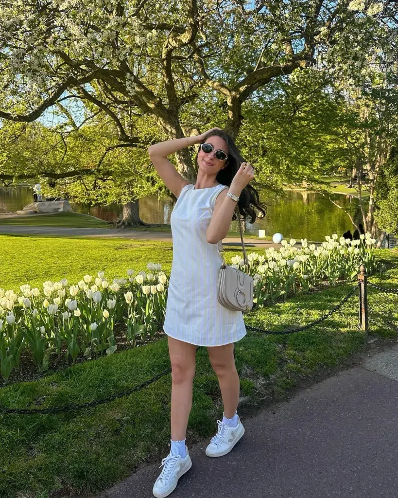 Petite woman standing on a park path in spring, wearing a pale pastel vertical-striped sleeveless mini shift dress with a boat neckline, white ruffle-cuff ankle socks, and white low-top sneakers, carrying a small taupe saddle crossbody bag, with round sunglasses and a thin gold necklace, tulips and blossoming trees behind her.