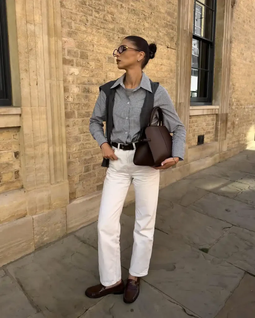 A woman standing on stone pavement in warm afternoon light against a honey-toned brick building facade, wearing a dark charcoal structured sleeveless vest layered over a blue and white fine vertical-stripe button-front shirt with a spread collar, paired with high-waisted straight-leg white jeans with a slim black leather belt visible at the waist, dark chocolate brown penny loafers, and carrying a structured dark chocolate brown leather top-handle bag, with hair in a tight bun and round tortoiseshell sunglasses.