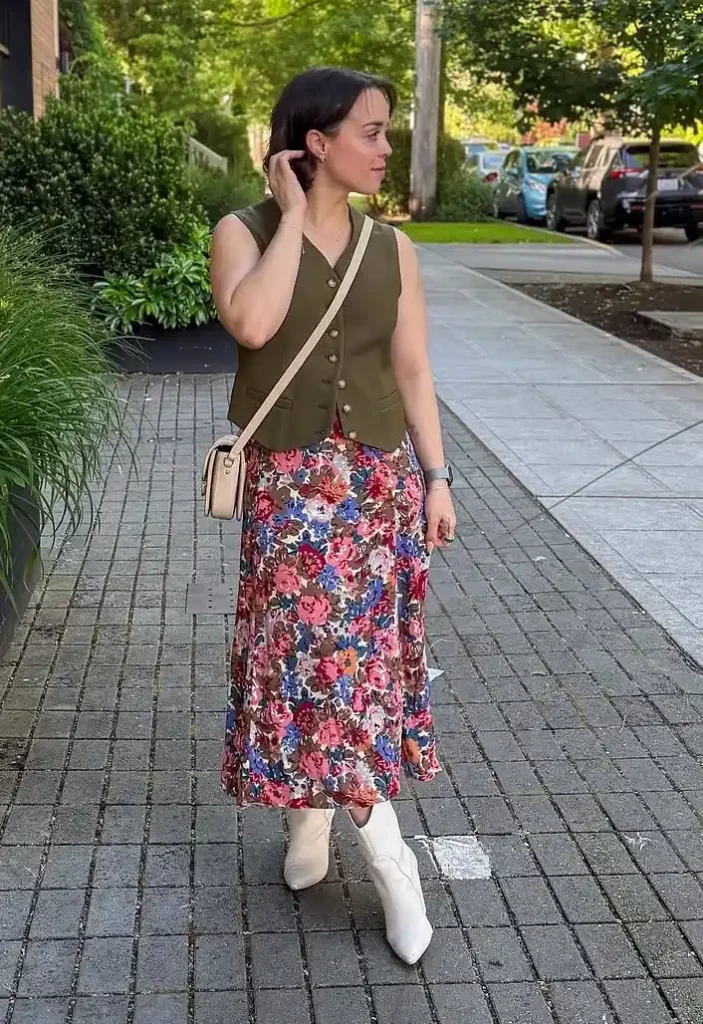 A petite woman walking on a cobblestone city sidewalk, wearing an olive green structured button-front waistcoat with gold button detailing over a bold vintage floral midi skirt in pink, red, blue, and brown tones, paired with white pointed-toe ankle boots and a small cream crossbody bag, with lush green trees in the background.