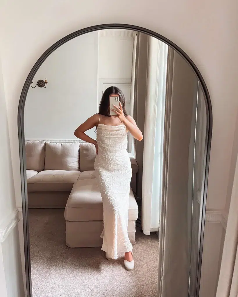Petite woman taking a mirror selfie in an elegant living room, wearing a floor-length white textured slip maxi dress with a cowl neckline and thin straps, paired with white espadrille flats, standing in front of a cream sofa beneath a black arched mirror.