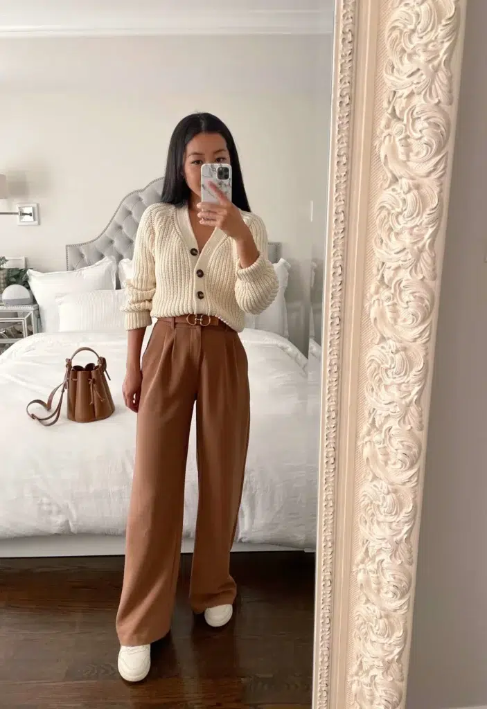 Petite woman in a mirror selfie wearing a cream chunky knit cardigan with brown buttons over high-waisted wide-leg caramel trousers with a slim gold-hardware belt and white sneakers, with a tan bucket bag in the background — a casual warm-toned petite interview outfit for creative or startup environments