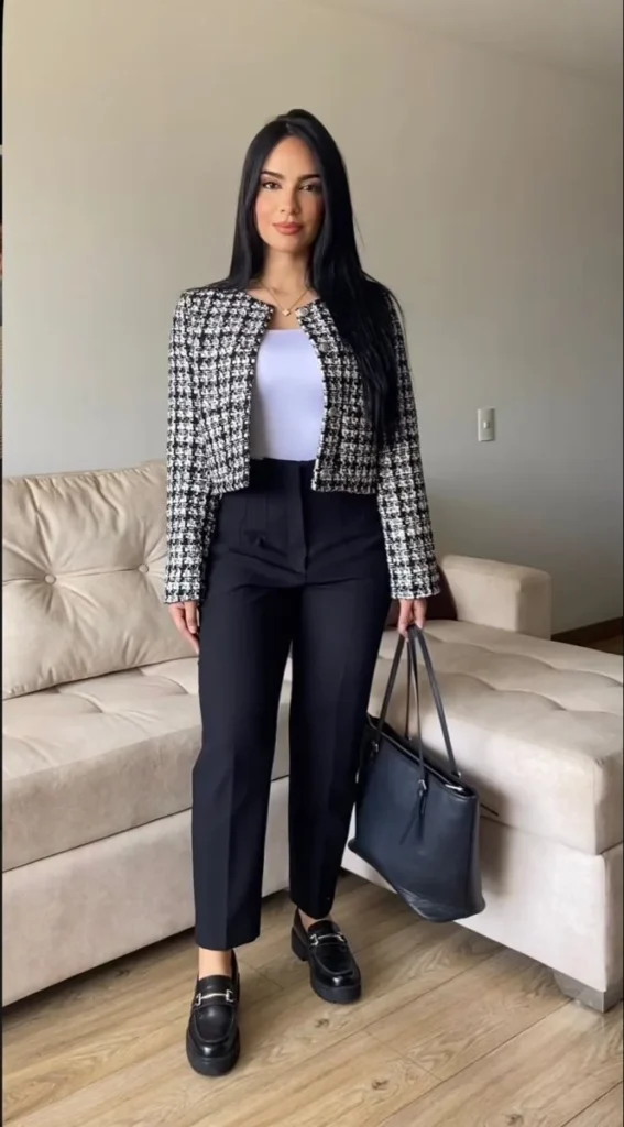 Petite woman wearing a cropped collarless black and white houndstooth tweed jacket with gold buttons over a light lavender fitted tank top and high-waisted black straight-leg ankle trousers, with chunky black platform loafers with gold hardware and a large black structured tote bag — a modern smart-casual petite interview outfit