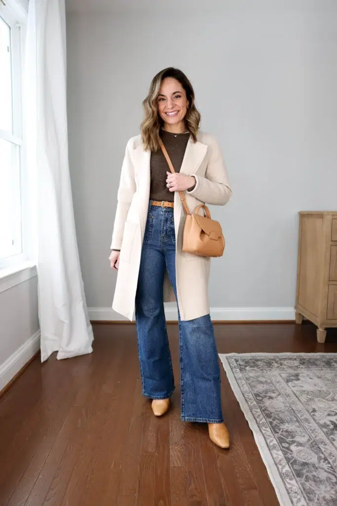 Petite woman in dark wash wide-leg flare jeans and a long open cream wool-blend coat, worn over a fitted brown crew-neck sweater with a cognac leather belt visible at the waist, styled with tan ankle boots and a structured camel top-handle bag.