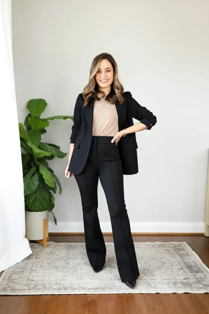 Petite woman wearing an oversized longline black blazer open over a camel fitted knit top and high-waisted black flare trousers with black pointed-toe heels, standing in a bright minimal room — a modern relaxed-professional petite interview outfit with flare pants