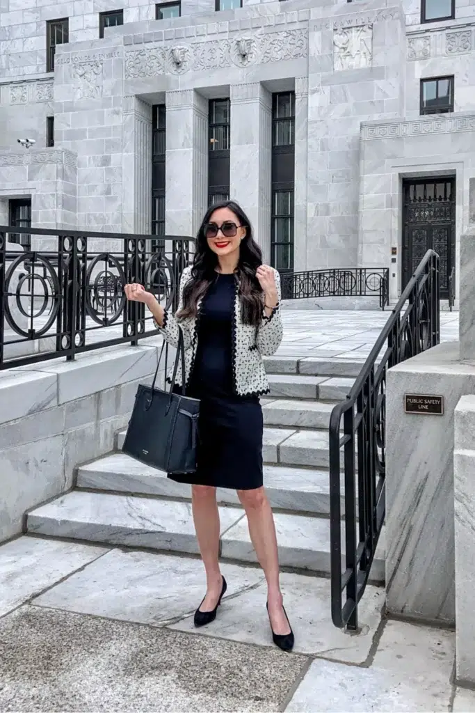 Petite woman in a cream and black scalloped-trim tweed-style cardigan jacket layered over a fitted navy sleeveless sheath dress, black pointed-toe heels, a large black structured tote, and bold red lipstick, standing on marble steps outside an official building — a fashion-forward petite interview outfit with a statement jacket