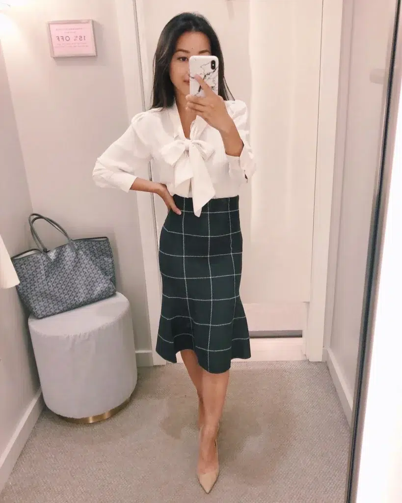 Petite woman in a fitting room mirror selfie wearing a white bow-tie puff sleeve blouse tucked into a dark green windowpane plaid midi skirt with a fluted ruffle hem, with nude pointed-toe heels and a grey patterned tote bag visible in the background — a polished petite interview outfit mixing two statement pieces