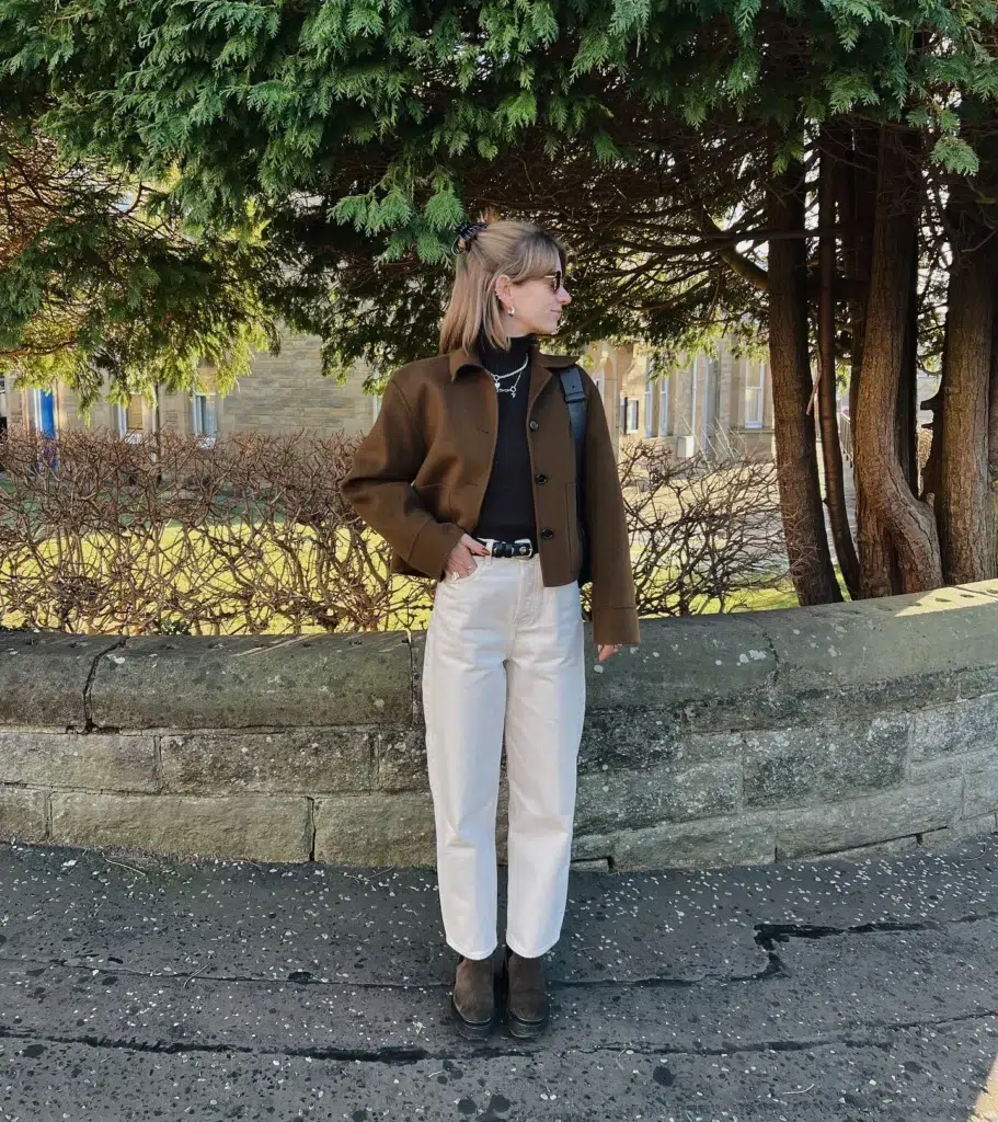 Petite woman with light brown shoulder-length hair half-up in a black claw clip standing outdoors against a curved stone wall with tall conifer trees and a stone building visible behind her, wearing a structured cropped olive tobacco brown wool chore jacket with button front and wide collar over a fitted black ribbed turtleneck with layered delicate silver necklaces, cream ecru barrel-leg jeans with a slim black leather belt with silver buckle at the waist, dark brown suede chunky ankle boots, small round tinted sunglasses, and a black backpack worn on one shoulder.