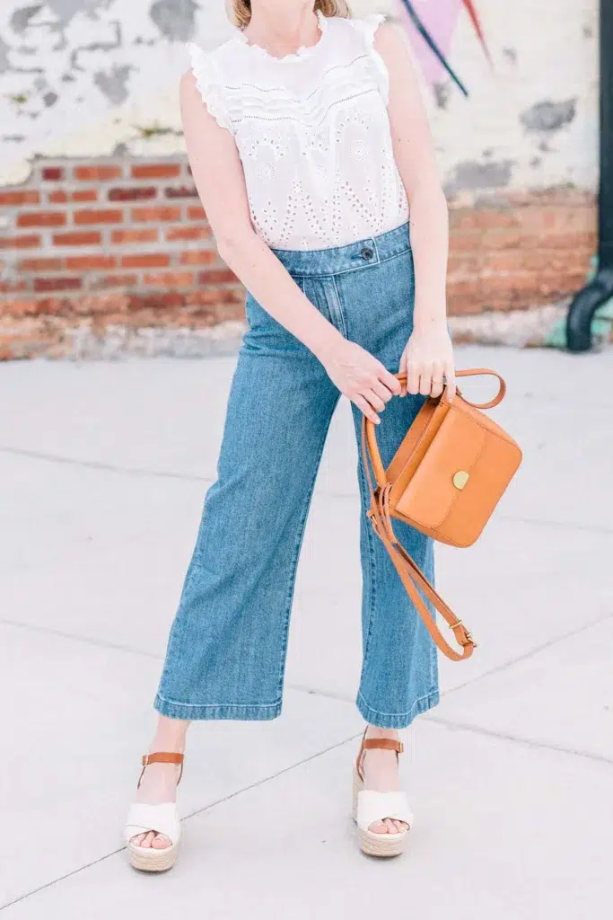 Petite woman styling high-rise wide-leg cropped jeans with a tucked-in white ruffled eyelet blouse, white espadrille platform sandals, and a tan leather crossbody bag.
