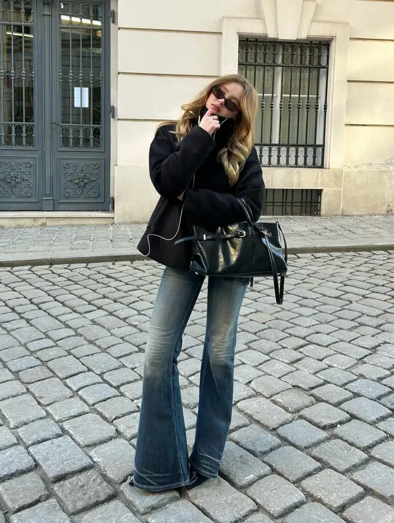 Petite woman in vintage-washed medium-dark flare jeans with an uneven, aged denim wash, wearing an oversized boxy black coat or jacket with a subtle white edge trim detail, paired with a large crinkled patent black leather shoulder bag with gold buckle hardware and dark pointed-toe boots on European cobblestones.