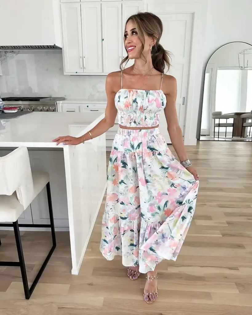 Petite woman in a matching watercolor floral two-piece co-ord set with a smocked spaghetti strap crop top and a tiered maxi skirt in white with pastel pink, lavender, teal, and coral flowers, wearing clear crystal-embellished heeled sandals and layered delicate gold necklaces, standing in a modern white kitchen.