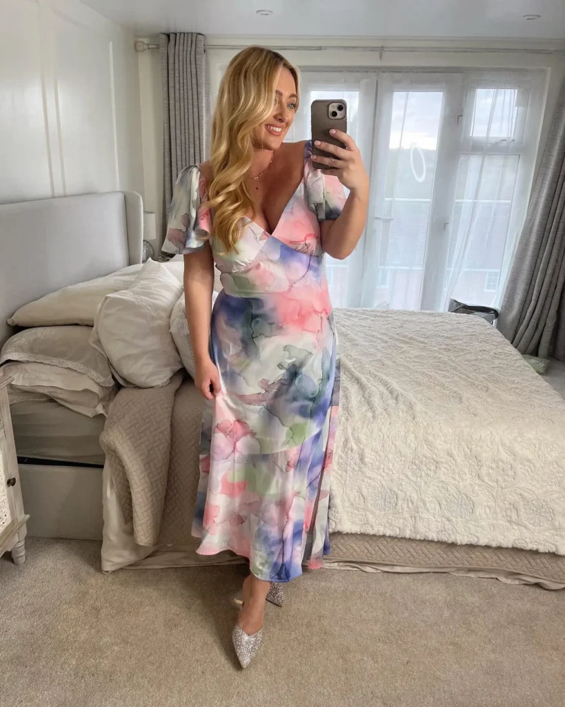 Curvy petite woman taking a bedroom mirror selfie wearing a floor-length wrap maxi dress in a soft watercolor abstract print with pink, lavender, sage green, and white tones, featuring a deep V-neckline, crossover bodice with a defined waist, and short flutter cap sleeves, paired with crystal-embellished pointed-toe heeled mules, a delicate gold necklace, and voluminous blonde wavy hair worn loose.