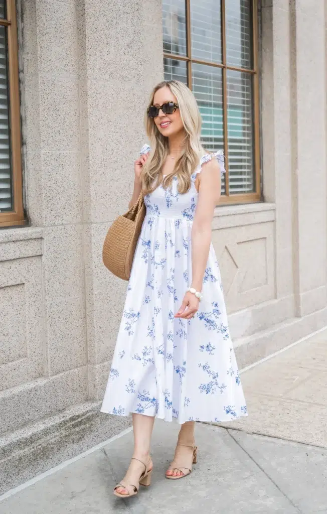 A petite woman in a white and blue floral smocked maxi dress with flutter ruffle sleeves, paired with nude block heel sandals, a round woven straw bag, and tortoiseshell sunglasses, photographed on an urban sidewalk against a stone building.