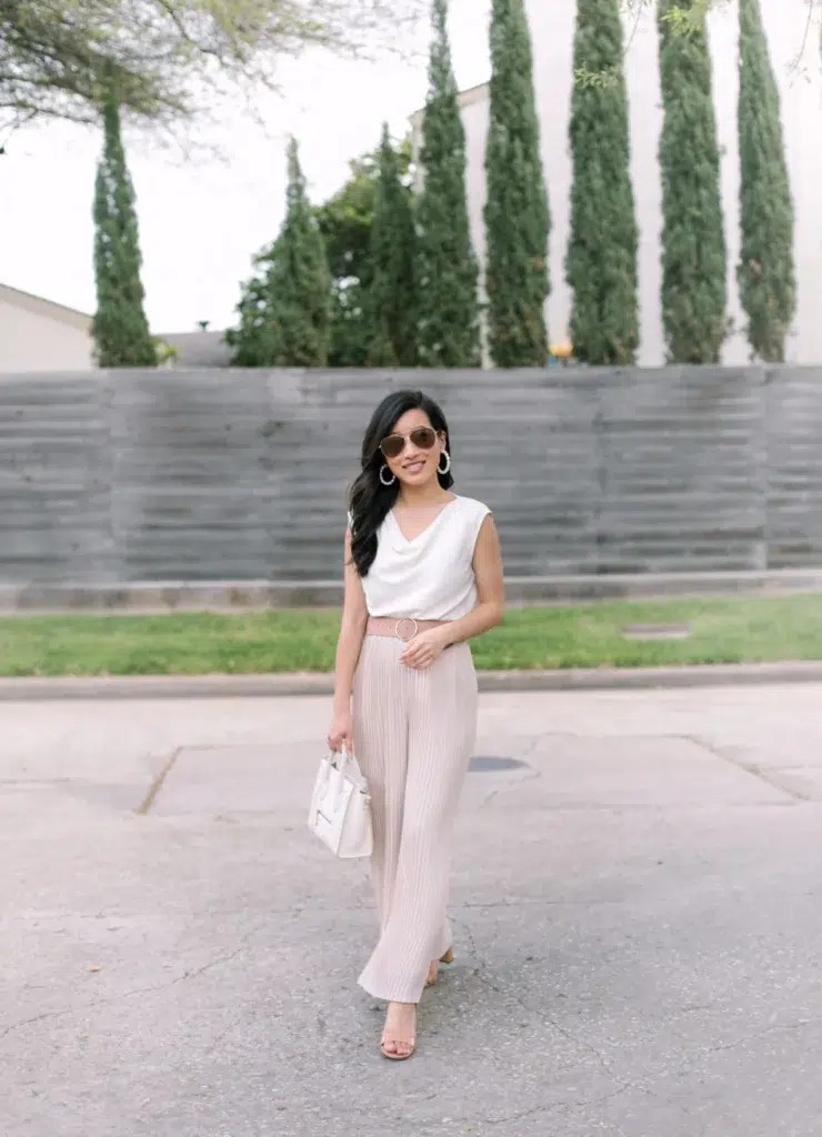 Petite Asian woman with long dark wavy hair and aviator sunglasses smiling outdoors on a sidewalk with tall cypress trees and a grey wooden fence behind her, wearing a white sleeveless cowl-drape top tucked into high-rise blush pink vertically ribbed wide-leg trousers with a matching blush circle ring-buckle belt, nude strappy low block-heel sandals, large pearl hoop earrings, and holding a small white structured satchel bag with gold hardware.