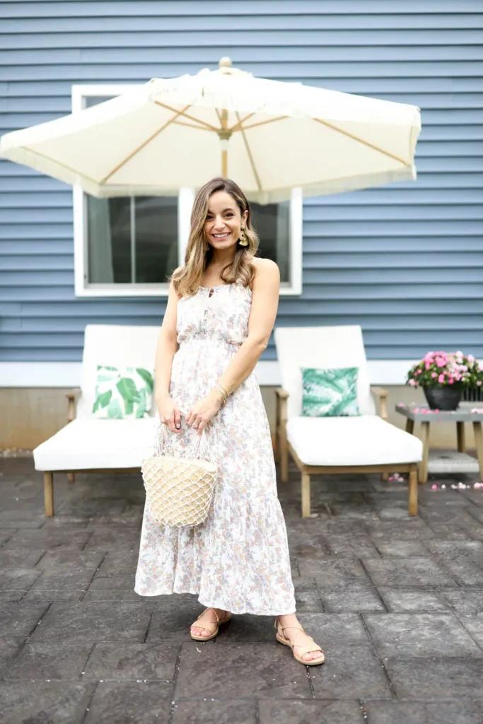 A petite woman wearing a white maxi dress with a small-scale muted floral print, spaghetti straps, a smocked blouson waist, and a tiered flowy skirt, styled with tan lace-up flat sandals, a round woven rattan bag with fringe trim, and oversized gold tassel earrings, photographed on a patio with lounge chairs and a cream umbrella in the background.