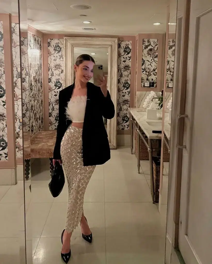 Petite woman in a white feather-trim strapless crop top, a champagne gold sequin fitted midi skirt, and an open black oversized blazer, styled with black patent pointed-toe pumps and a small black bag, taking a mirror selfie in an ornate powder room with floral wallpaper.