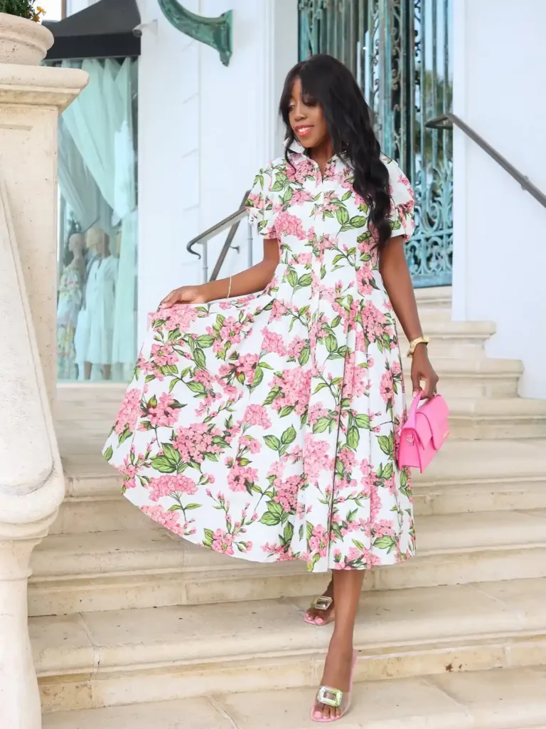 Woman in a white full-swing midi dress covered in large pink hydrangea floral clusters with bold green leaves, featuring a pointed shirt collar, short flutter sleeves, and a button-front bodice, wearing clear heeled mule sandals with a green jeweled buckle detail and carrying a small bright pink structured mini top-handle bag with gold hardware, posing on marble steps in front of an elegant white building with decorative green metalwork.