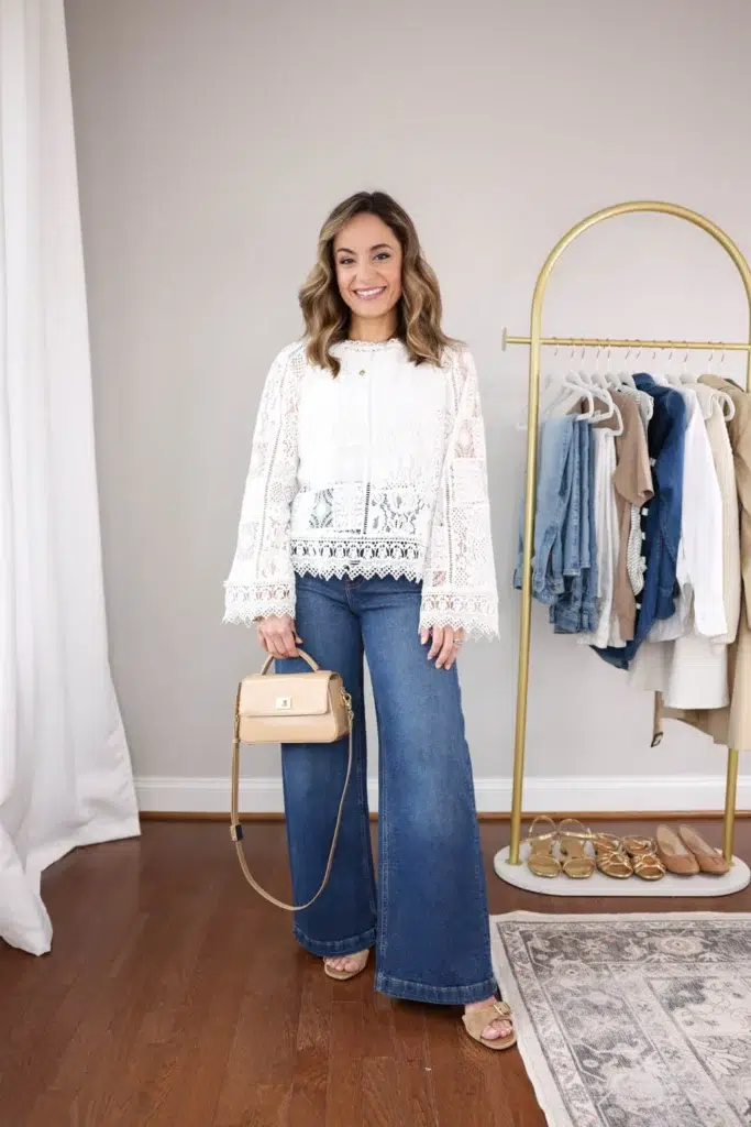 Petite woman in a white lace bell-sleeve blouse with a scalloped hem, high-rise dark wash wide leg jeans, nude block heel sandals, and a small structured tan top-handle bag