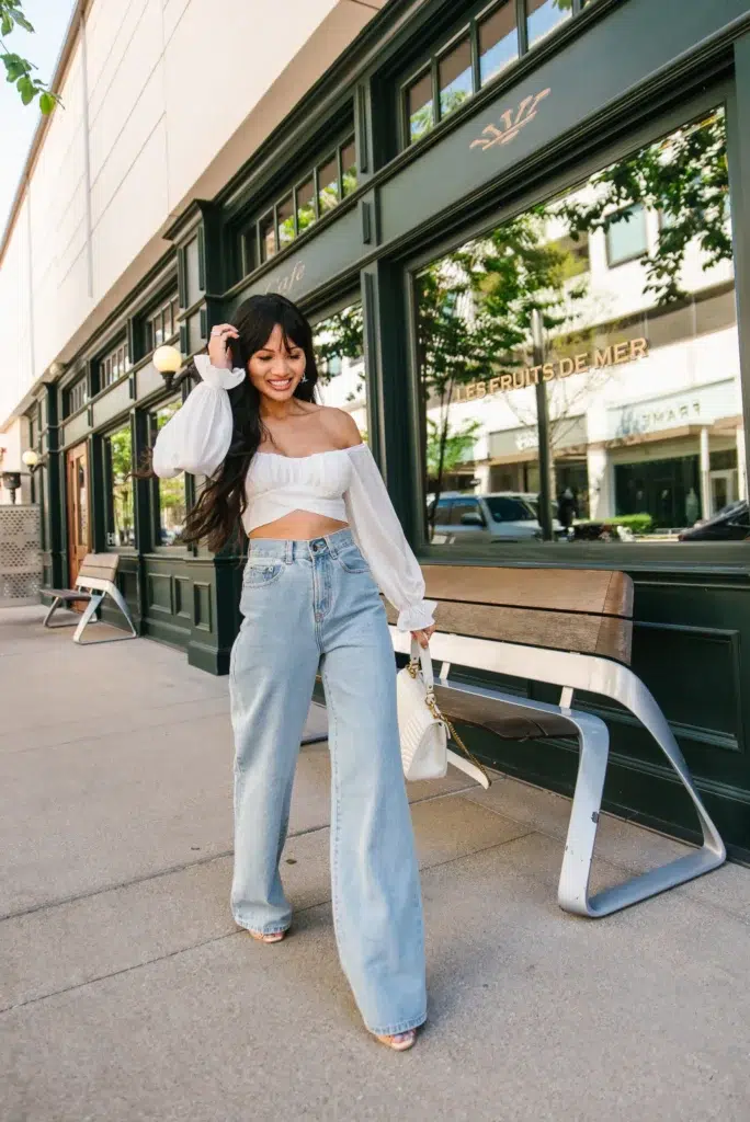 Petite woman in high-rise light wash wide-leg jeans and a white off-shoulder long-sleeve crop top with billowy sleeves and a front twist detail, carrying a white quilted chain-strap shoulder bag, standing on a city sidewalk in front of a green-framed café storefront.