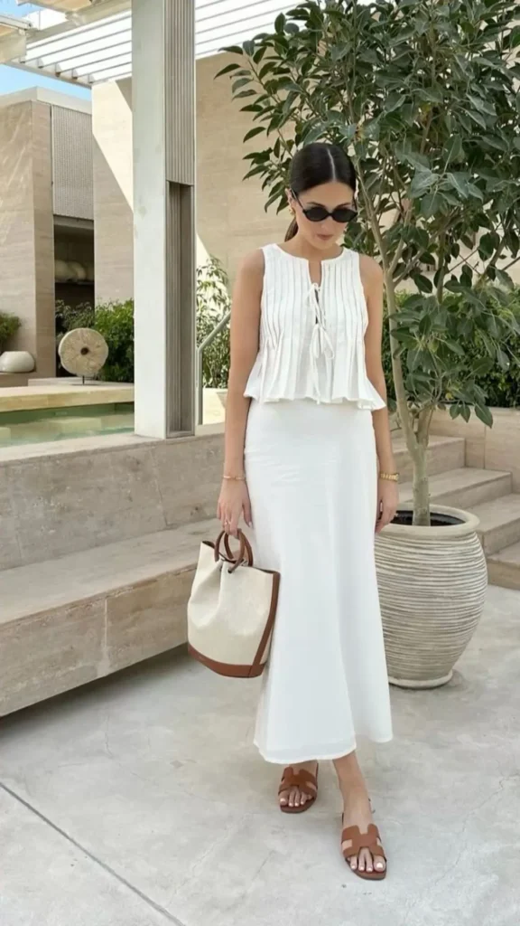 Petite woman in a white sleeveless accordion-pleated ruffle-hem top with a tie-front neckline paired with a fitted white column maxi skirt, wearing cognac brown H-strap flat sandals and carrying a structured canvas and cognac leather tote bag, standing on an outdoor travertine terrace with stone steps and an olive tree in a large ribbed ceramic pot in the background.