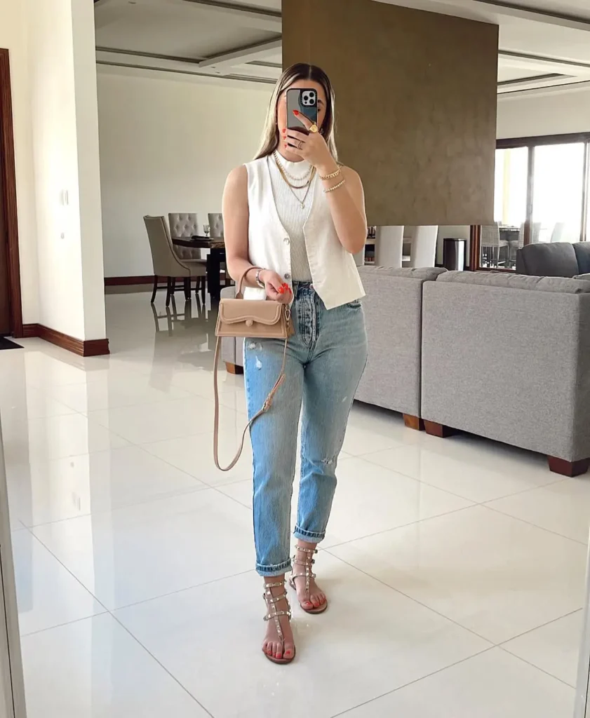 Woman taking a mirror selfie in light wash distressed mom jeans with a rolled cuff hem, a white structured button-front vest layered over a ribbed fitted top, studded flat sandals in nude, and a beige structured crossbody bag, with layered gold necklaces, indoors in a modern home