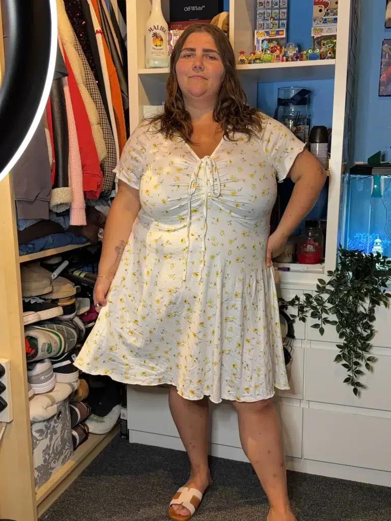 Curvy petite woman standing indoors wearing a white short-sleeve mini dress with a small yellow sunflower print and a ruched tie-front center detail, paired with white and tan flat slide sandals, with a cluttered closet and shelving unit visible in the background.