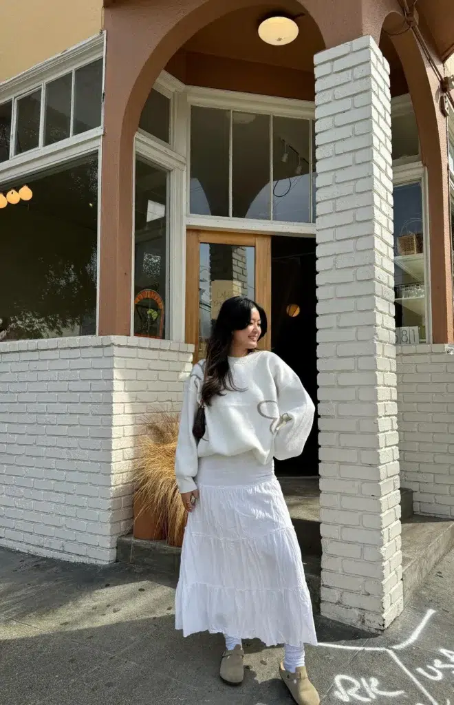 Petite woman standing outside a café on a brick corner, wearing an ivory oversized sweater with a tonal swirl detail, a white tiered cotton maxi skirt, slouchy white socks, and taupe suede clog mules, with a small crossbody bag, painted white brick behind her.