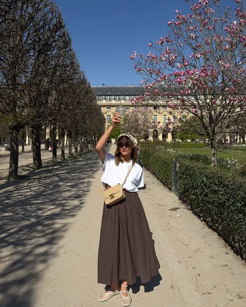 Petite woman standing on a tree-lined garden path in Paris, wearing a white tee knotted at the waist over a full chocolate-brown maxi skirt, a plaid headscarf, round sunglasses, sheer floral flats, and a small cream crossbody bag, holding an iced drink, with magnolia blossoms behind her.
