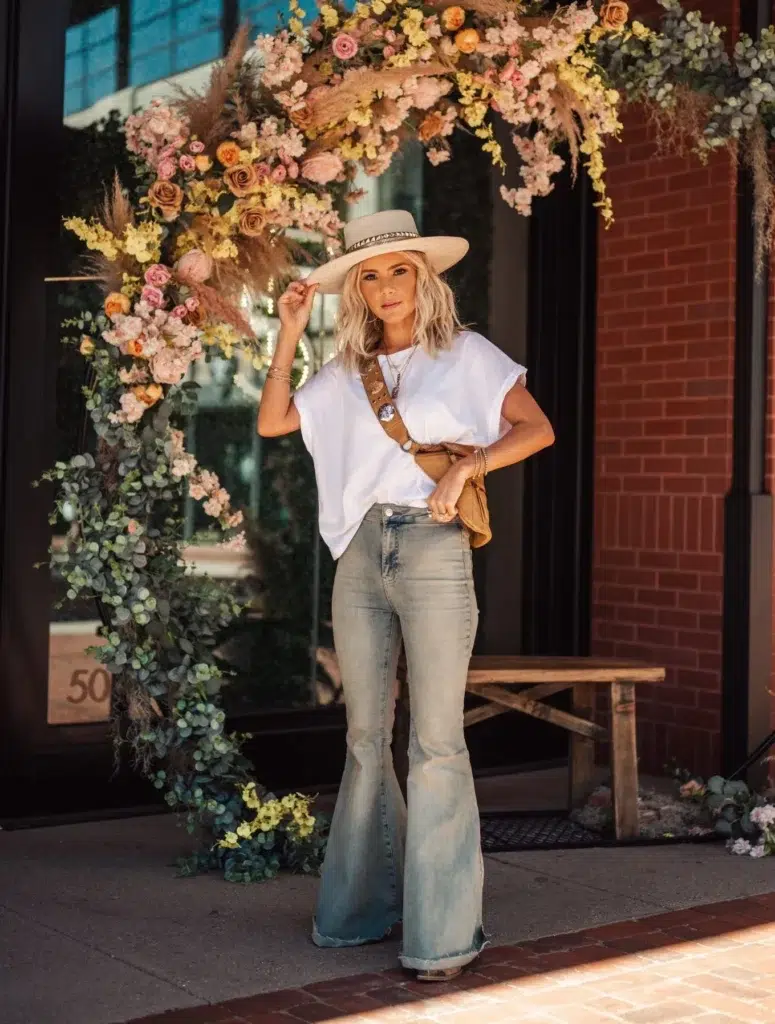 Petite woman in very light wash faded flare jeans with a raw hem, wearing a white boxy short-sleeve tee and a wide-brim structured straw hat with a decorative band, styled with a tan suede belt bag on a tooled leather strap, layered gold necklaces, and stacked bracelets.