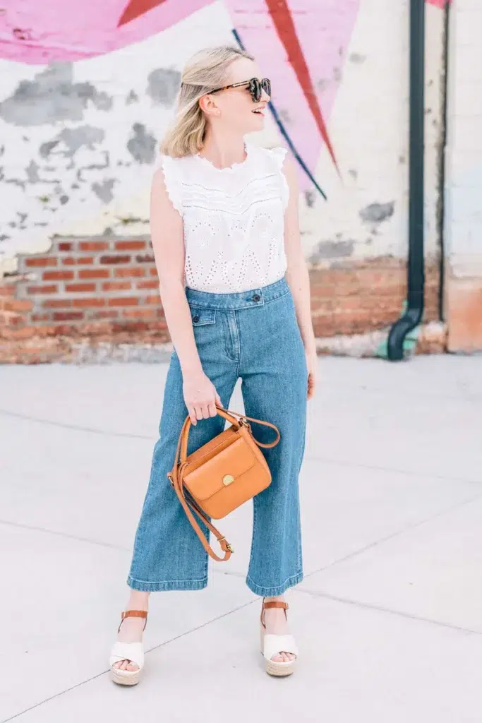 Petite woman wearing high-rise wide-leg cropped jeans with a tucked-in white eyelet blouse, tan leather crossbody bag, and espadrille wedge sandals showing how to wear wide leg jeans petite with balanced proportions