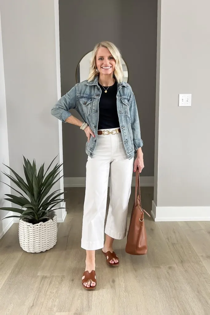 Petite woman wearing white high-rise wide-leg cropped pants with a tucked-in black top, brown belt, cropped denim jacket hitting above the hip, and brown slide sandals showing how to layer over wide-leg pants without hiding the waistline