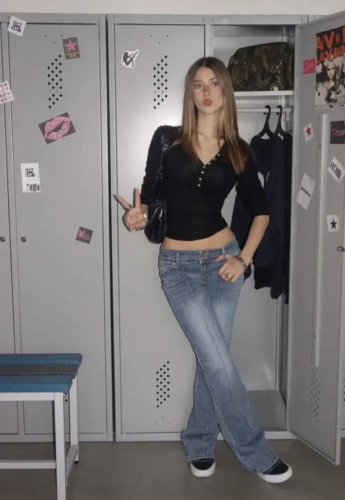 Young woman in low-rise light wash flare jeans with a faded wash and slight boot cut shape, wearing a fitted black long-sleeve top with a V-neckline and button placket detail, styled with black and white low-top sneakers, a small black patent mini shoulder bag, and stacked silver rings and bracelets in a sticker-covered locker room setting.