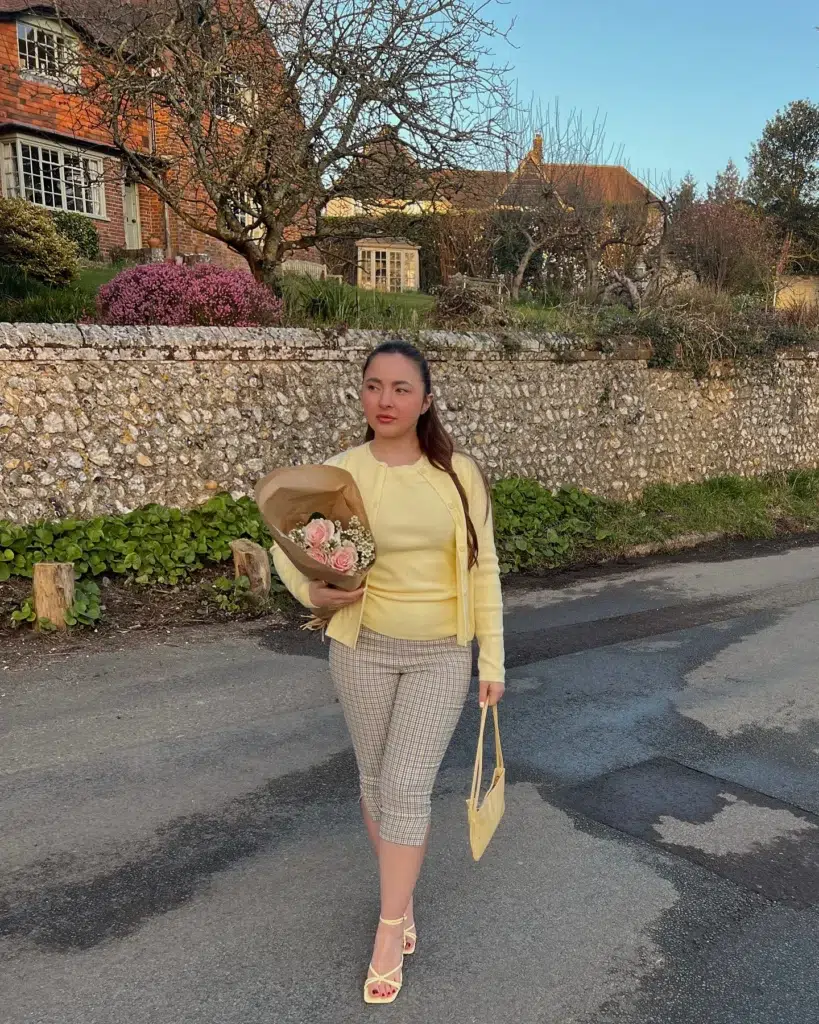 Petite woman walking on a country road holding pink roses, wearing a butter-yellow knit twin set with an open cardigan, cropped beige gingham capri pants, strappy yellow heels, and a small yellow shoulder bag, with a flint wall and brick cottage behind her.