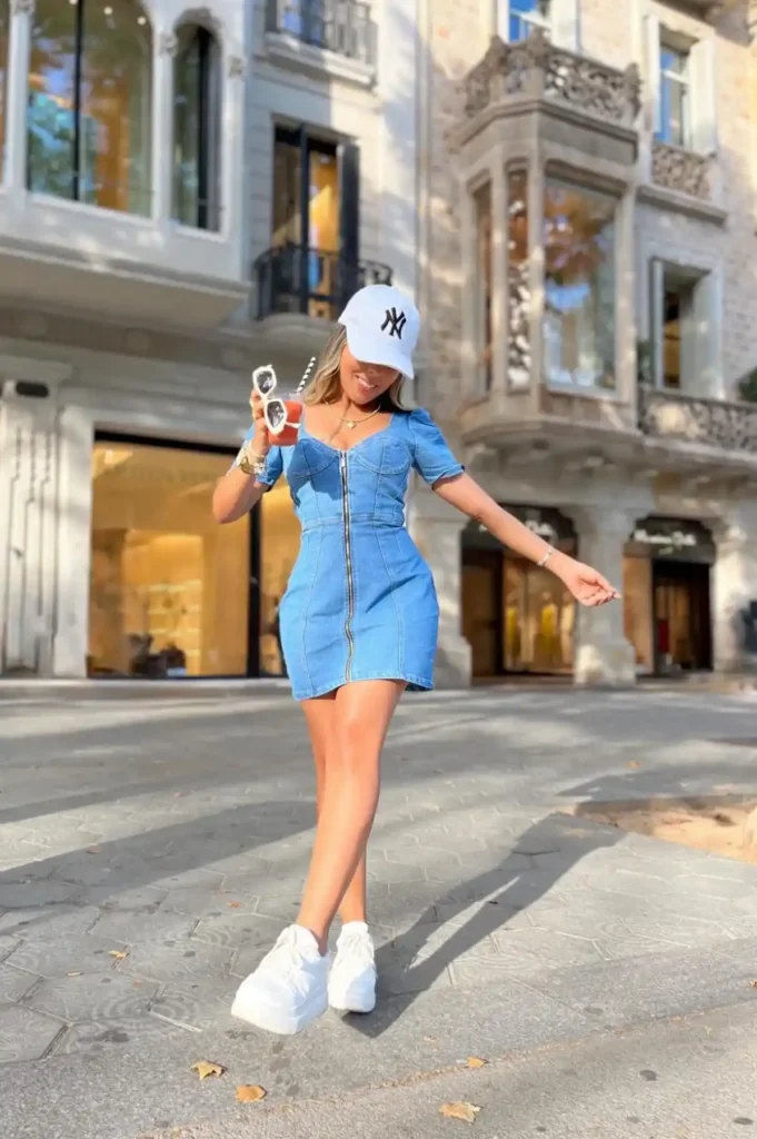 Young woman in a medium blue wash fitted zip-front denim mini dress with corset-style vertical seam lines and short puff sleeves, styled with a white baseball cap with a black logo, white chunky platform sneakers with white ankle socks, layered gold chain necklaces, and a gold watch, holding an iced drink and a pair of white sunglasses while standing on a sunlit European city street with ornate stone buildings in the background.