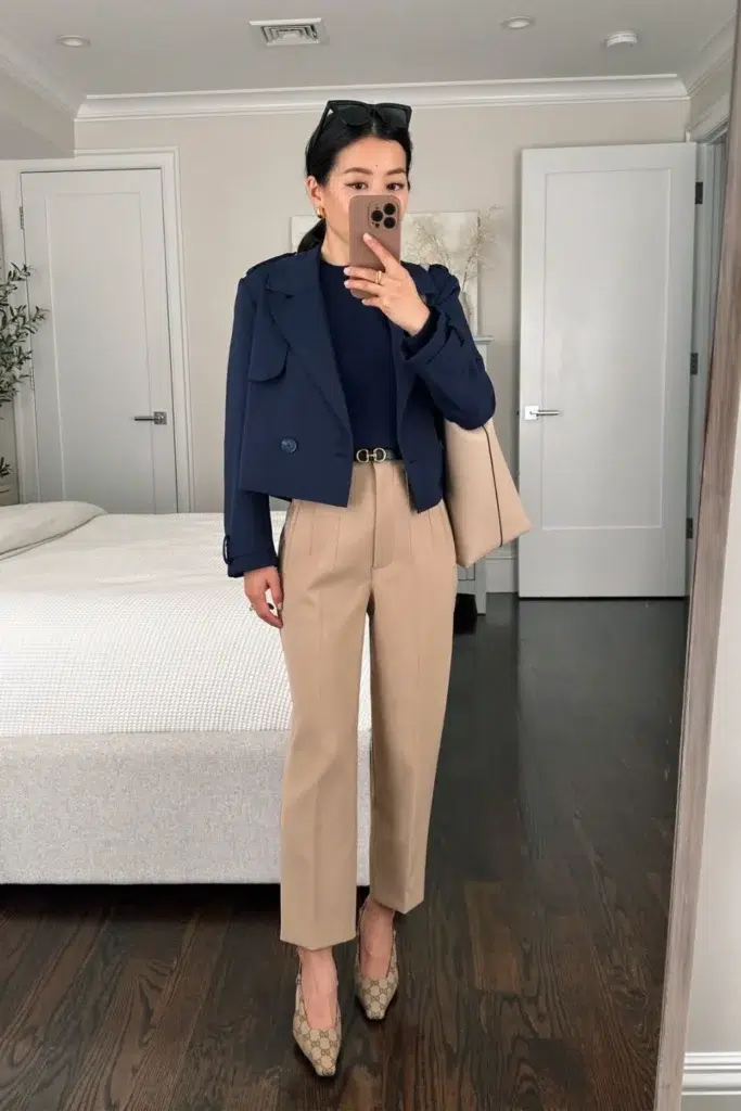 A petite woman in a cropped navy blazer ending at the hip bone over a navy top and beige pleated trousers, with a thin belt visible at the waist and patterned pointed-toe pumps, showing how a shorter blazer length keeps the torso-to-leg ratio balanced on a petite frame.