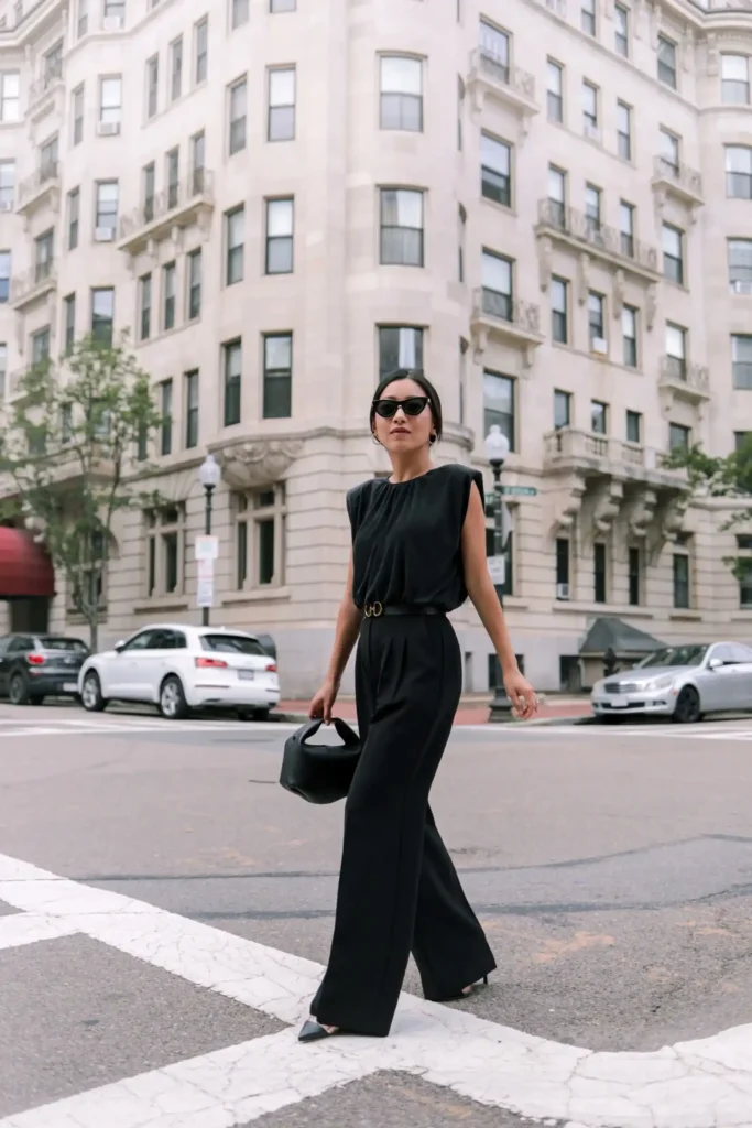 A petite woman in an all-black work outfit with a sleeveless top tucked into wide-leg trousers, a black belt with gold hardware at the waist, black pointed-toe heels, and a black handbag, showing how keeping top and bottom in the same color family creates one unbroken vertical line from shoulder to shoe.