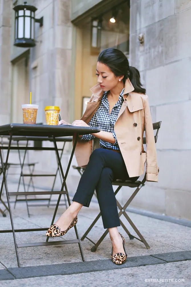 A seated woman in navy cropped work trousers and leopard-print pointed-toe flats with a low vamp and no ankle strap, showing how a pointed-toe shoe shape extends the leg line without any horizontal break at the ankle.