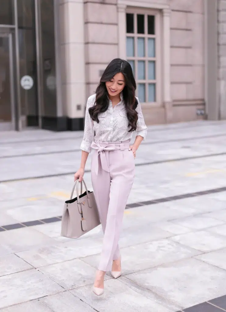 A petite woman with a printed button-up blouse fully tucked into high-waisted lilac paperbag trousers with a bow-tie waist, paired with nude pointed-toe pumps and a taupe structured handbag, showing how tucking the blouse defines the waistline and gives the legs more visible length.