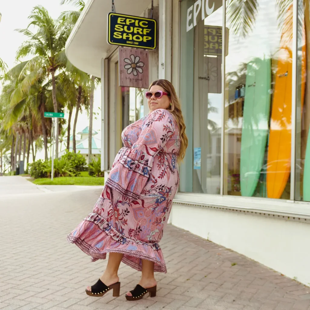 Petite plus-size woman with long wavy blonde hair wearing a long-sleeve tiered boho maxi dress in pink with a mixed paisley and floral print in red, blue, and lavender, featuring ruffle tier panels and a decorative border-print hem, styled with black suede block heel wooden mule clogs with gold stud trim detail, pink cat-eye sunglasses, and colorful dangle earrings, caught mid-turn on a brick paver sidewalk outside a coastal surf shop with palm trees and colorful surfboards visible through the storefront window behind her.