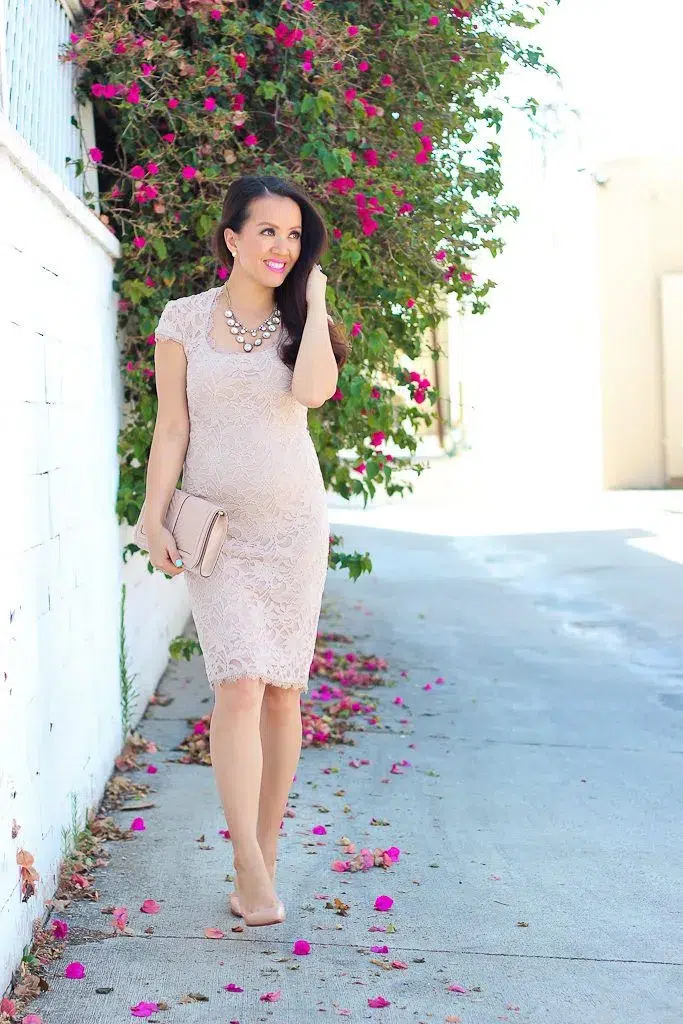 Petite pregnant woman with long dark wavy hair, wearing an all-over blush pink lace fitted sheath dress with cap sleeves, a scoop neckline, and a scalloped lace hem that falls just above the knee, revealing a clearly visible baby bump, wearing nude pointed-toe heels and carrying a blush pink envelope clutch, with a multi-strand crystal and pearl bubble statement necklace at the collarbone and a bright pink lip, smiling and walking along a sunlit whitewashed alleyway with a wall of vivid magenta bougainvillea flowers and green foliage in the background.