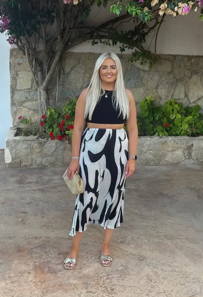 Petite woman in a fitted black sleeveless crop top and high-waisted abstract black and white bold-print pleated midi skirt, paired with gold metallic knot-detail flat slide sandals and a small woven straw clutch with gold hardware, standing outdoors against a stone wall with flowering plants.
