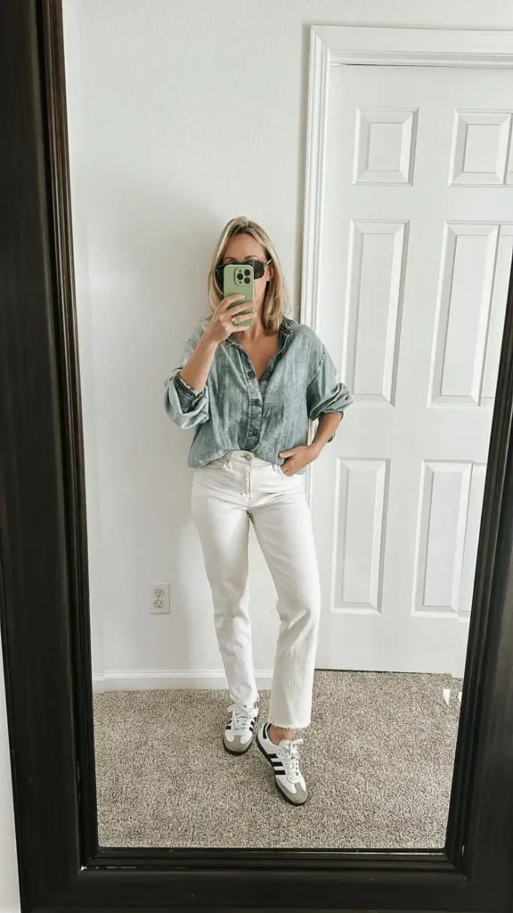 Petite woman with chin-length blonde hair taking a mirror selfie in a neutral-toned bedroom with beige carpet and a white panelled door, wearing white high-rise straight-leg ankle-crop denim jeans with a raw frayed hem, an oversized acid-wash faded blue-grey chambray button-down shirt with sleeves rolled to the elbow and a loose half-tuck at the front waistband, and white and black stripe low-profile sneakers in a classic Samba or Gazelle silhouette.