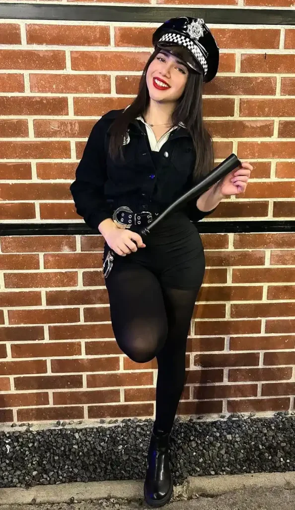 Petite woman with long dark hair and red lips wearing an all-black Halloween police costume outfit consisting of an oversized structured black jacket, white collared shirt underneath, black high-waisted mini shorts, a wide black belt with silver buckle, and black opaque tights, accessorized with a shiny black and white checkered police officer cap, holding a black prop baton, standing against a brick wall at night.