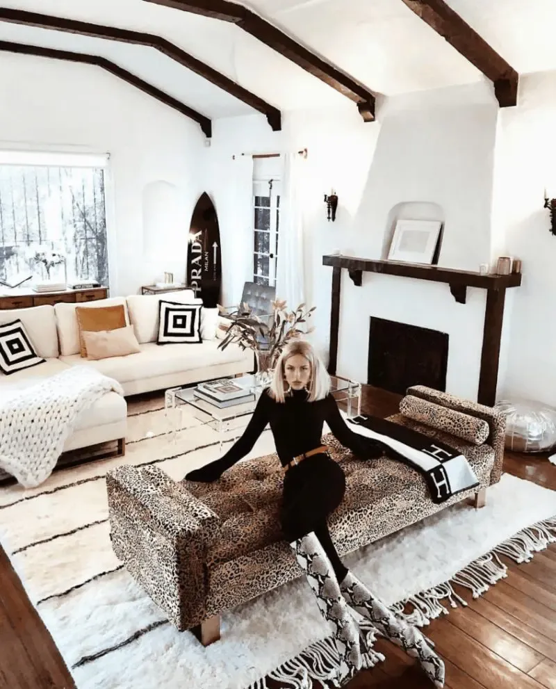 Petite woman with short blonde hair seated on a leopard-print tufted chaise longue, wearing an all-black fitted long-sleeve turtleneck top and black slim-fit jeans cinched with a thin cognac leather belt, paired with snake-print ankle boots, photographed in a Spanish-style white plaster living room with dark wood ceiling beams, a cream sectional sofa, and a white shag rug.