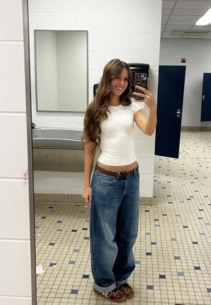 Petite woman taking a bathroom mirror selfie wearing ultra-baggy medium-wash boyfriend jeans sitting low on the hip with a brown leather belt and a wide ankle cuff, paired with a fitted white ribbed crop baby tee, with leopard print mule slippers visible at the hem.