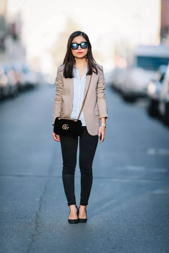 Petite woman with straight dark hair standing in the middle of a city street, wearing black skinny denim jeans cropped above the ankle, a pale light blue button-down shirt, and a fitted single-button beige blazer hitting at the hip, paired with black flat shoes and a small black velvet chain crossbody bag with gold logo hardware, wearing large mirrored cat-eye sunglasses and a silver watch.