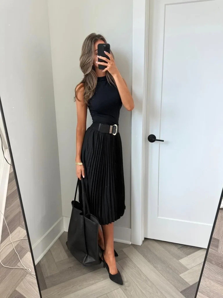 A petite woman in an all-black business casual outfit: a sleeveless mock-neck top, a belted pleated midi skirt, and black pointed heels, with a large black leather tote, photographed in a mirror selfie.