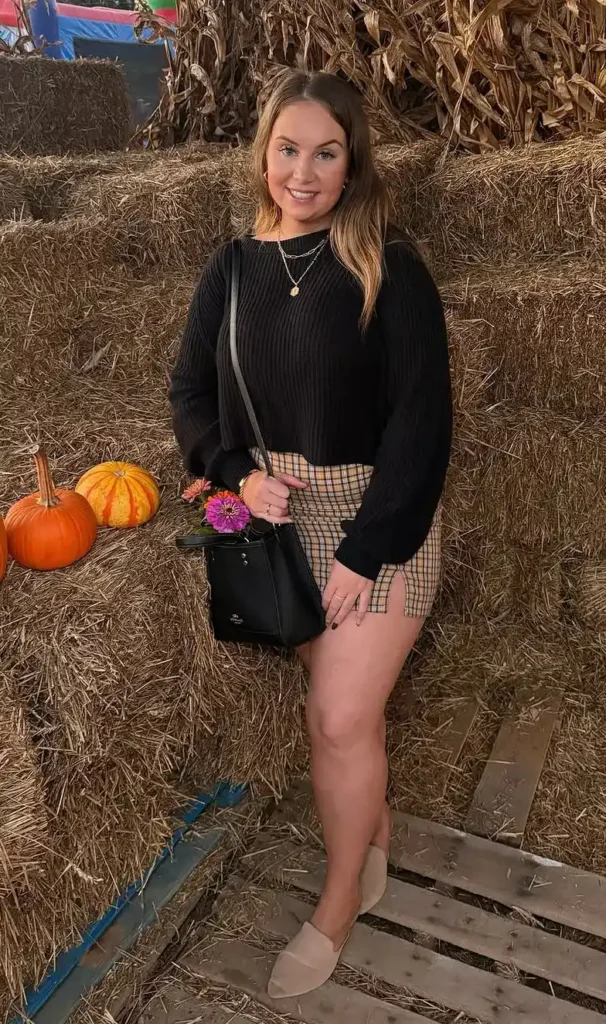 Petite woman in a black ribbed cropped long-sleeve sweater stopping precisely at the waistband of a high-waisted camel and dark plaid check mini skirt, wearing nude flat slip-on mules, carrying a small black structured bucket bag on a crossbody strap, with layered delicate gold necklaces, smiling and leaning lightly against stacked hay bales with orange pumpkins beside her.