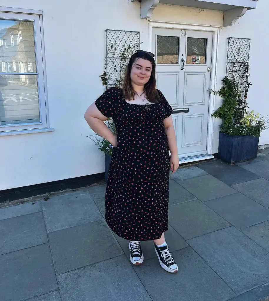 Petite plus-size young woman with straight medium-length brown hair and sunglasses pushed up on her head, smiling with one hand on her hip, wearing a black midi dress with a small scattered red ditsy floral print, a gathered square milkmaid-style neckline with a small center tie detail, short puff sleeves, an empire waistline, and a relaxed flowing midi skirt, paired with white ankle socks and black and white platform high-top lace-up sneakers with a gum sole, and a thin delicate layered necklace, standing on grey stone paving slabs in front of a white painted house with a white panelled front door numbered 50 and green climbing plants in grey planters on either side.