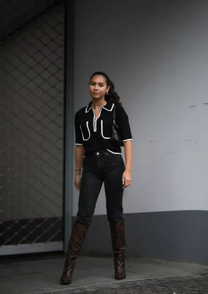 Petite woman with dark hair in a low ponytail standing outdoors against a grey concrete wall, wearing black high-waisted straight-leg denim jeans tucked over tall knee-high dark brown snake python-print leather boots, paired with a black short-sleeve polo-collar knit top with white contrast piping trim at the collar, pocket, and hem, accessorised with a small black crossbody bag, silver hoop earrings, a delicate chain necklace, and silver bracelets.